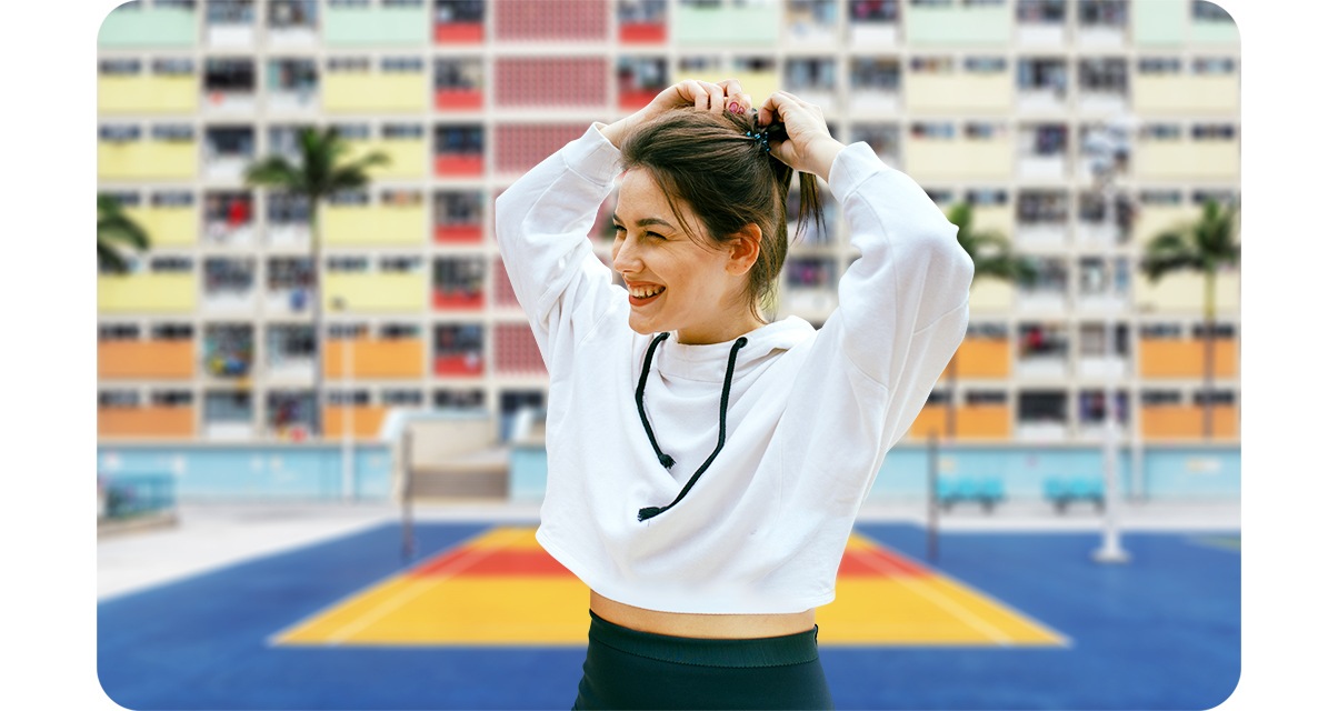 (Direita) O modo Retrato está desligado. Uma mulher está arrumando seu cabelo e sorrindo, olhando para a direita. Atrás dela está uma quadra de vôlei, apartamentos coloridos e árvores tropicais.