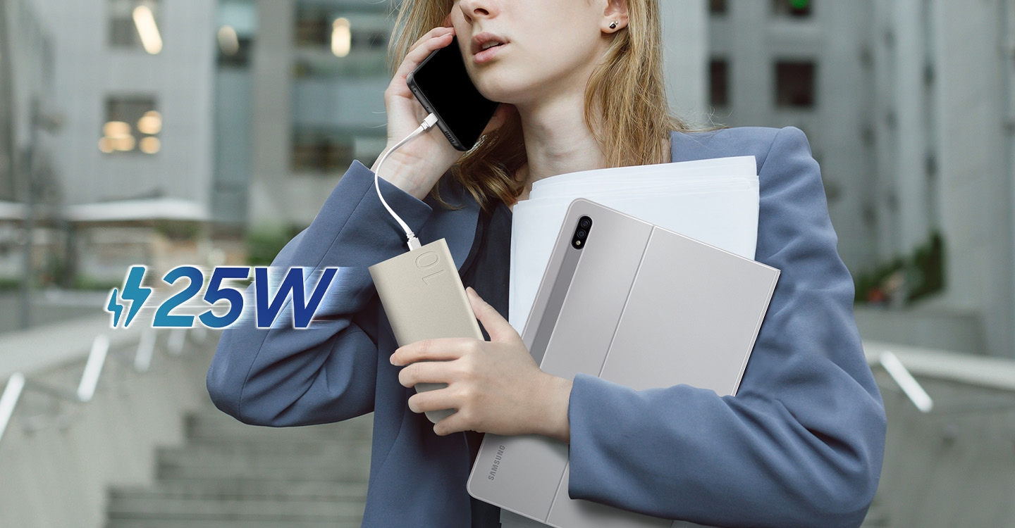 A woman in a business attire is in a call using Galaxy S22, which is connected to the battery pack via USB Type-C cable and charging in her hand, while also holding a Galaxy Tab S8+ and some papers. "25W" is shown on the left.