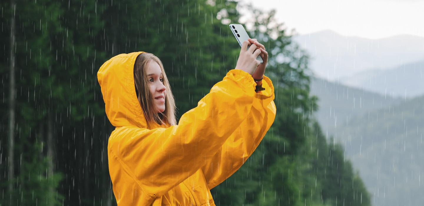 Uma pessoa sorrindo com uma capa de chuva amarela brilhante tirando uma selfie com um Galaxy A55 5G na chuva.