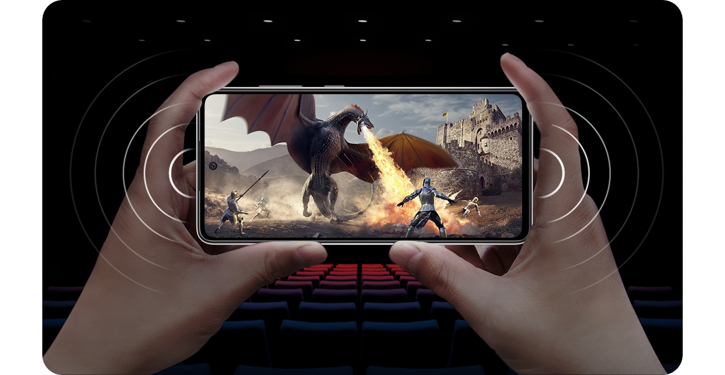 Uma pessoa segurando o Galaxy A52 no modo paisagem com uma cena de um cavaleiro lutando contra um drago que solta fogo e ondas sonoras vindo de ambos os lados do celular para demonstrar os alto-falantes estreo.