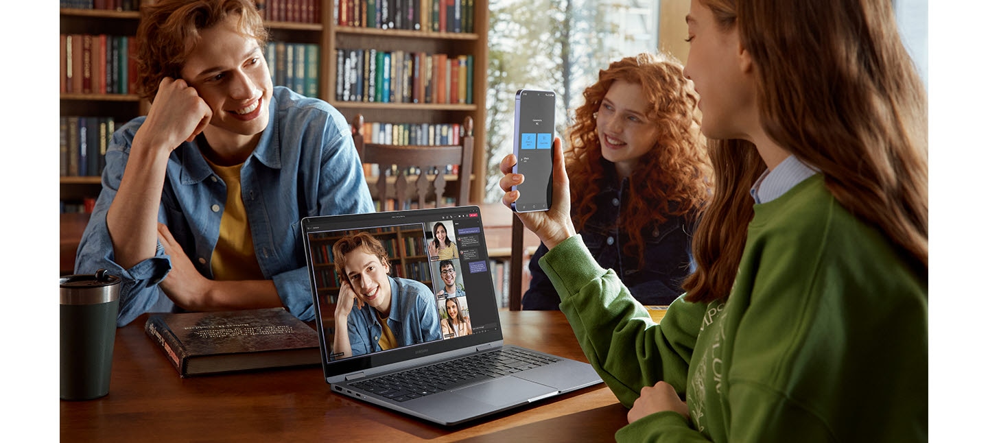 A young woman is using Galaxy S24 Plus together with two friends as a connected camera with Galaxy Book4 360 in Gray, placed on a table open and facing forward with Microsoft Teams group video call window and four participants shown onscreen.