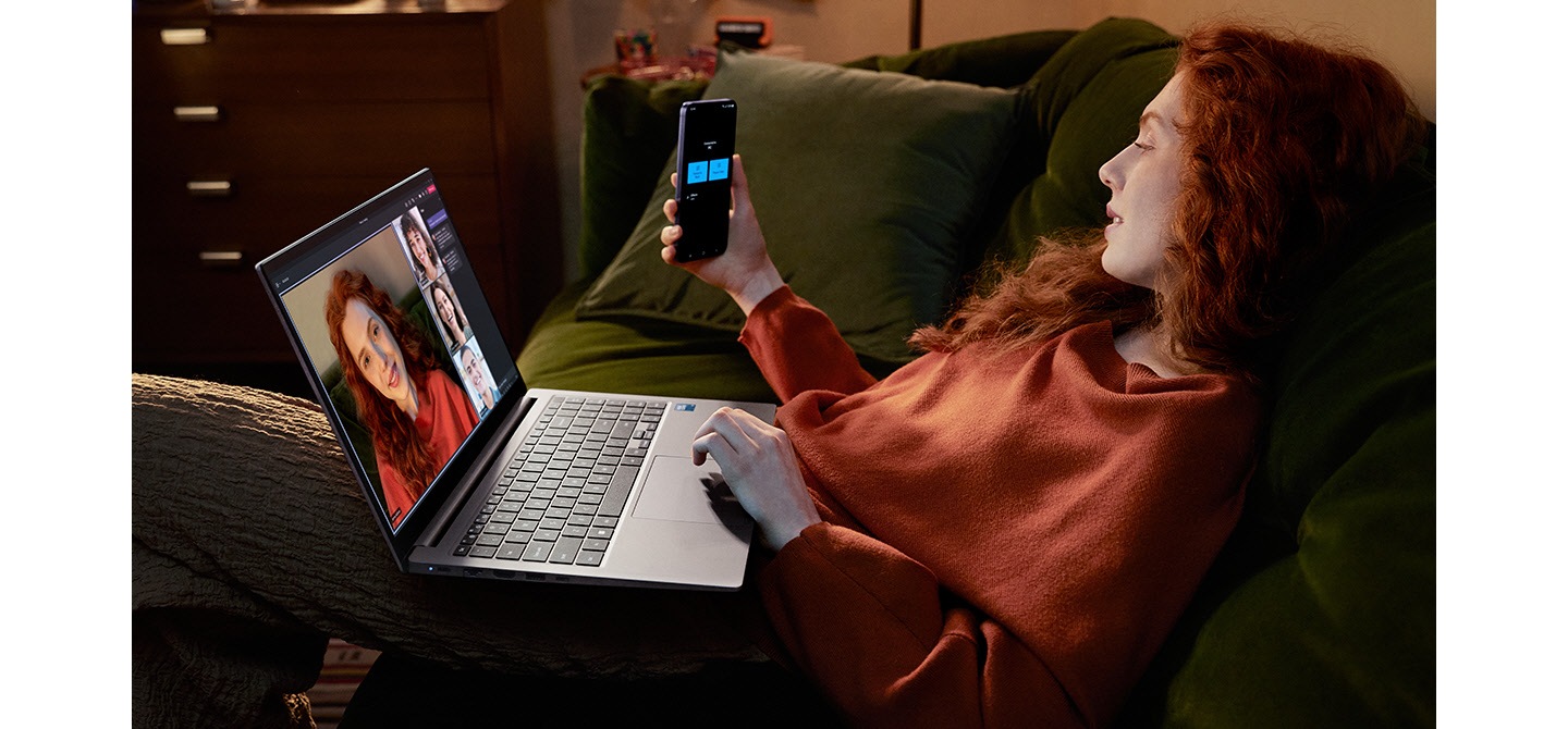 A young woman sitting on a sofa at home is using Galaxy S24 Plus as a connected camera with Galaxy Book4 in Gray to participate in a Microsoft Teams group video call with three other people shown onscreen.