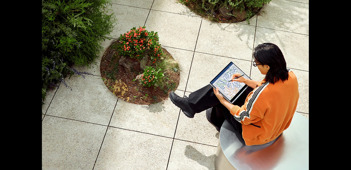 A young man sitting in a garden outdoors is using S Pen to edit a colorful image open in Goodnotes app onscreen on Galaxy Book4 Pro 360 in Platinum Silver, folded all the way backwards in tablet mode.