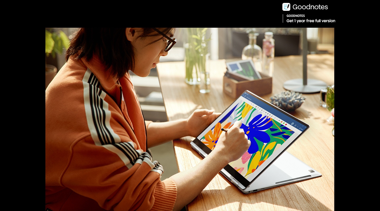 A young man is using S Pen to edit a colorful image open in Goodnotes app onscreen on Galaxy Book4 Pro 360 in Platinum Silver, folded backwards and placed on a table in a brightly lit studio. The onscreen image is clear and vibrant with improved visibility thanks to Vision Booster. Goodnotes logo with text 'Get 1 year free full version' is shown.