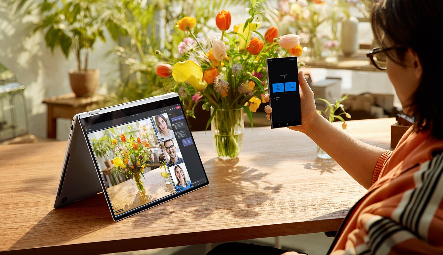 A person is using Galaxy S24 Ultra as a connected camera with Galaxy Book4 Pro 360 in Platinum Silver, placed on a table and folded like a tent with Microsoft Teams group video call window shown onscreen.