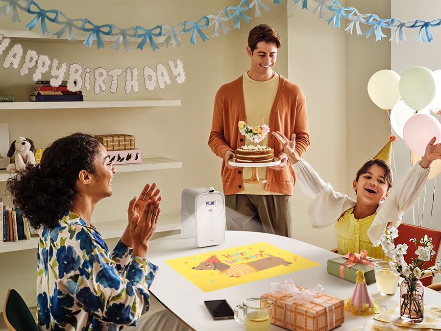 A happy family celebrates a birthday party. The Premiere 5 projects an image of a cartoon dog with the words Happy Birthday onto a cluttered table. With the Auto Scale feature, The Premiere 5 automatically resizes the image to be smaller, so that it doesn’t overlap with the clutter on the table.