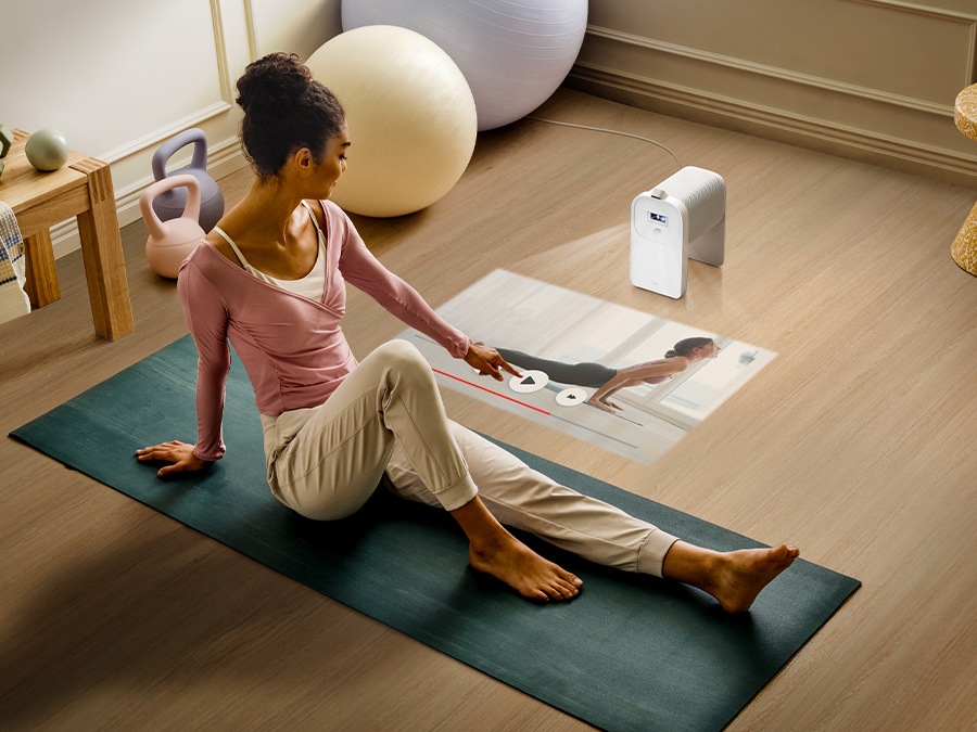 A woman is sitting on a yoga mat and watching a yoga tutorial video being projected onto the floor by The Premiere. She taps at the projected screen with her finger, causing the video to play. The yoga teacher in the video demonstrates a stretch.