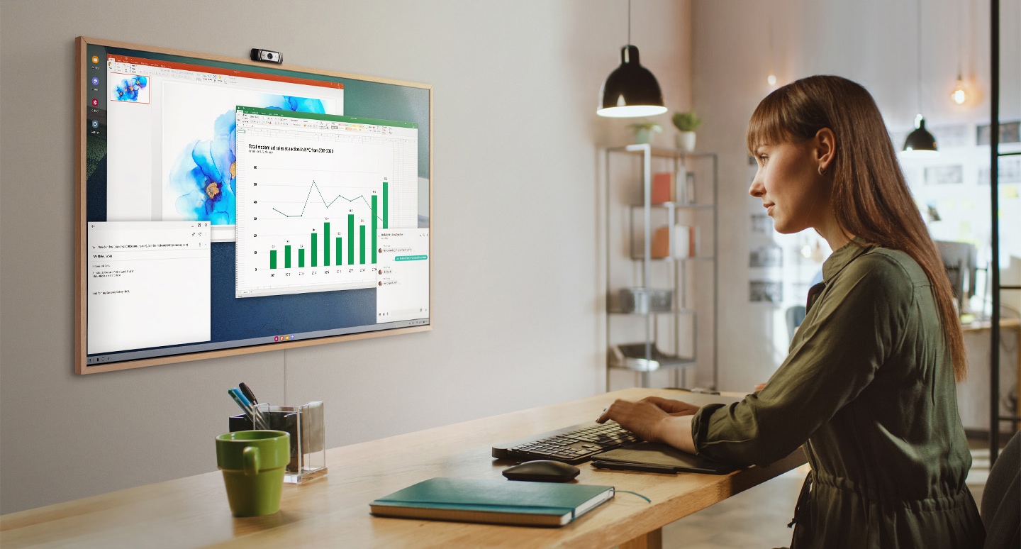 A woman is using her office PC to do work on her TV screen at home. On the screen of The Frame are various office productivity windows.