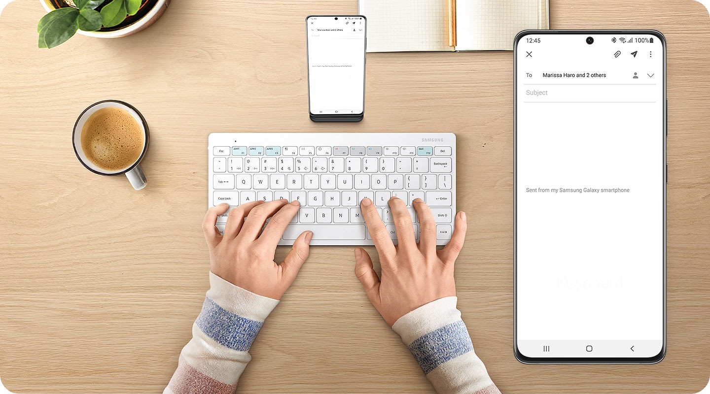 There is a white-colored Samsung Smart Keyboard Trio 500 on the desk and two hands are typing on the keyboard. In front of the keyboard, there is a smartphone placed on a phone holder and an email app is shown on the screen of the phone. On the right side, there is a close-up of what is on-screen on the phone.