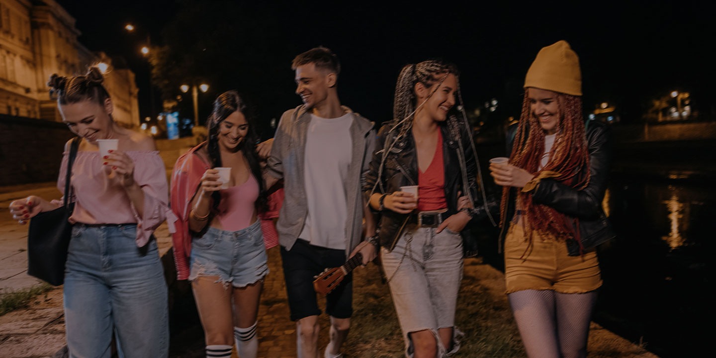 Cinco amigos, cuatro mujeres y un hombre, caminan junto al río de noche con bebidas en la mano bajo los faroles. Un deslizador vertical divide la imagen: a la izquierda, tonos apagados; a la derecha, colores brillantes que muestran detalles más claros, como un gorro amarillo, jeans rotos y reflejos en el agua.