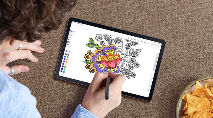 Con el S Pen, una persona está coloreando la imagen intrincada de una flor en la aplicación PENUP en un dispositivo Galaxy Tab S6 Lite mientras está sentada en un piso alfombrado con refrigerios a un lado.