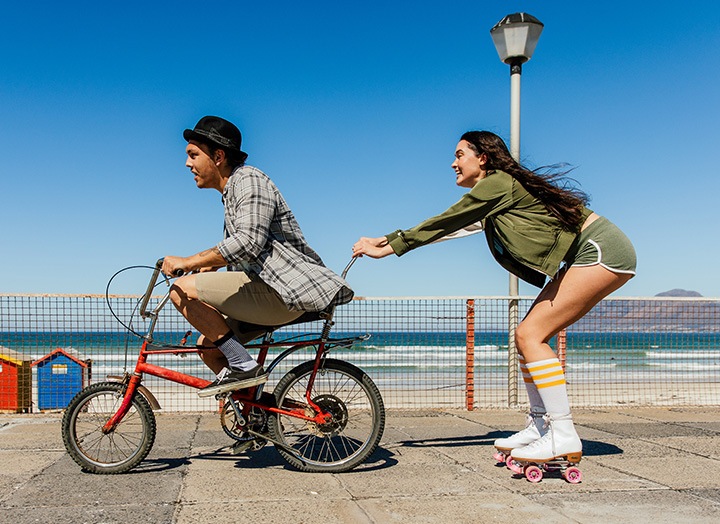 Una pantalla dividida con una línea en el centro muestra a un hombre y una mujer cerca de la playa. A la izquierda, que aparece ligeramente borrosa, el hombre monta en bicicleta. A la derecha, nítida y clara, la mujer se agarra a la bicicleta del hombre con unos patines, lo que pone de relieve la función OIS del Galaxy A26 5G.