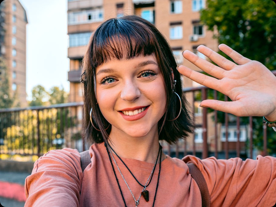 Una persona con una camisa de durazno y varios collares se toma una selfie al aire libre. Resalta la capacidad de la cámara frontal para tomar selfies impresionantes.