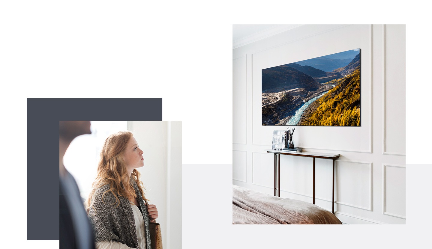 A woman and a man in a stylish white room look at a TV with a landscape background onscreen. The TV is on a white wall above a small narrow console that has a few ornaments on it.