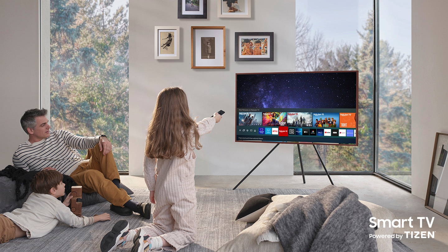 A father and his two children are enjoying The Frame which is mounted to Studio Stand. On the screen is the Universal Guide user interface menu which the girl is controlling with One Remote. The Smart TV Powered by Tizen logo is visible.
