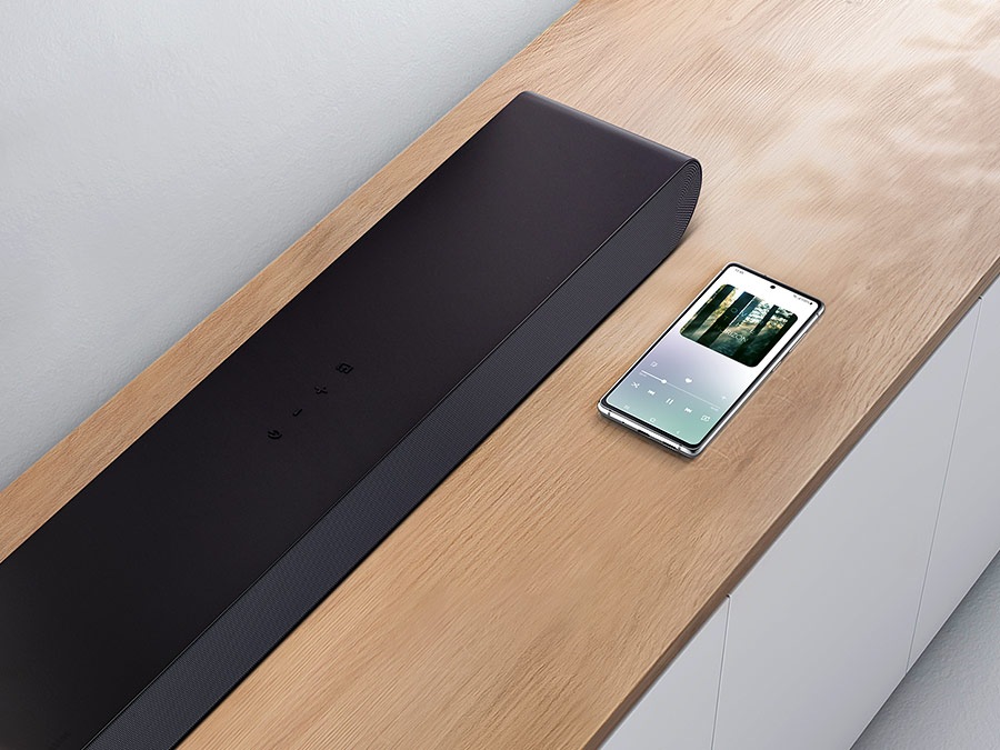A smartphone which is showing music screen, sits next to Samsung S50B soundbar demonstrating Bluetooth connectivity for smartphone music playback.
