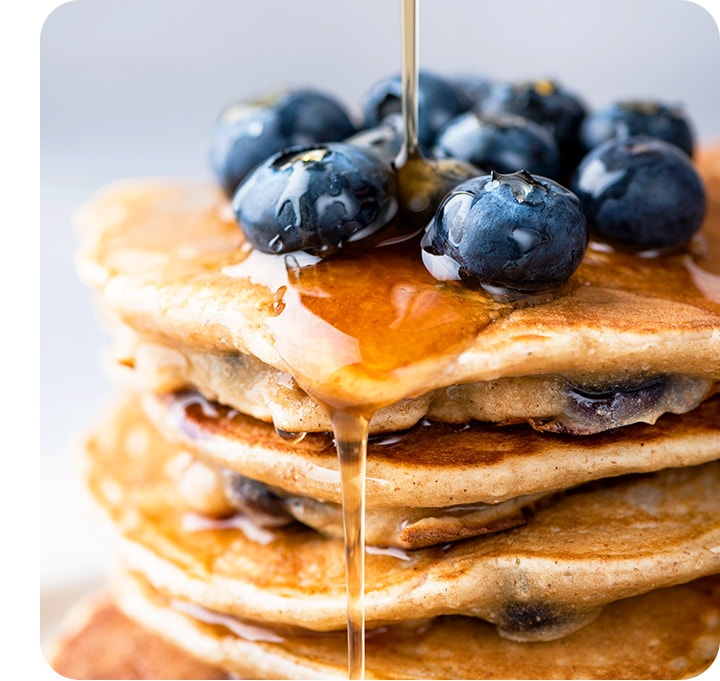 Syrup being poured onto a stack of pancakes with blueberries on top. The details can be seen clearly.
