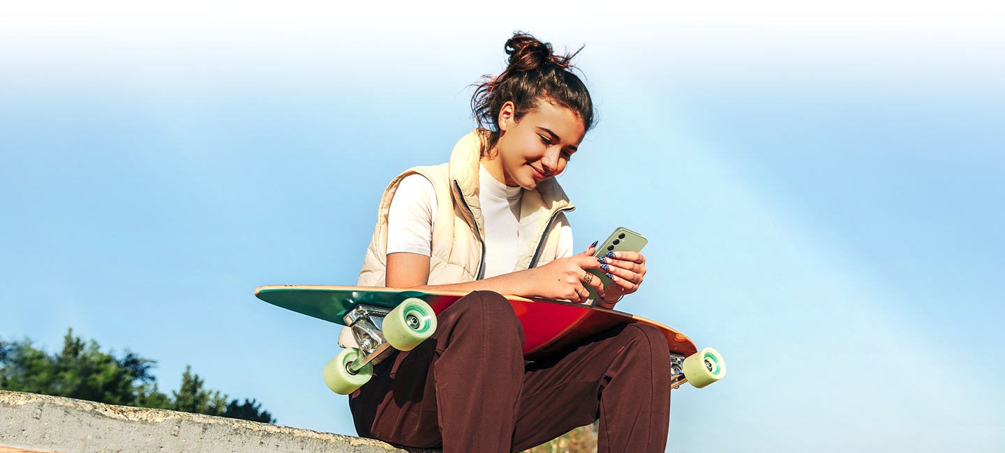 Sedící žena s longboardem na klíně sleduje displej telefonu Galaxy A54 5G na přímém slunečním světle.
