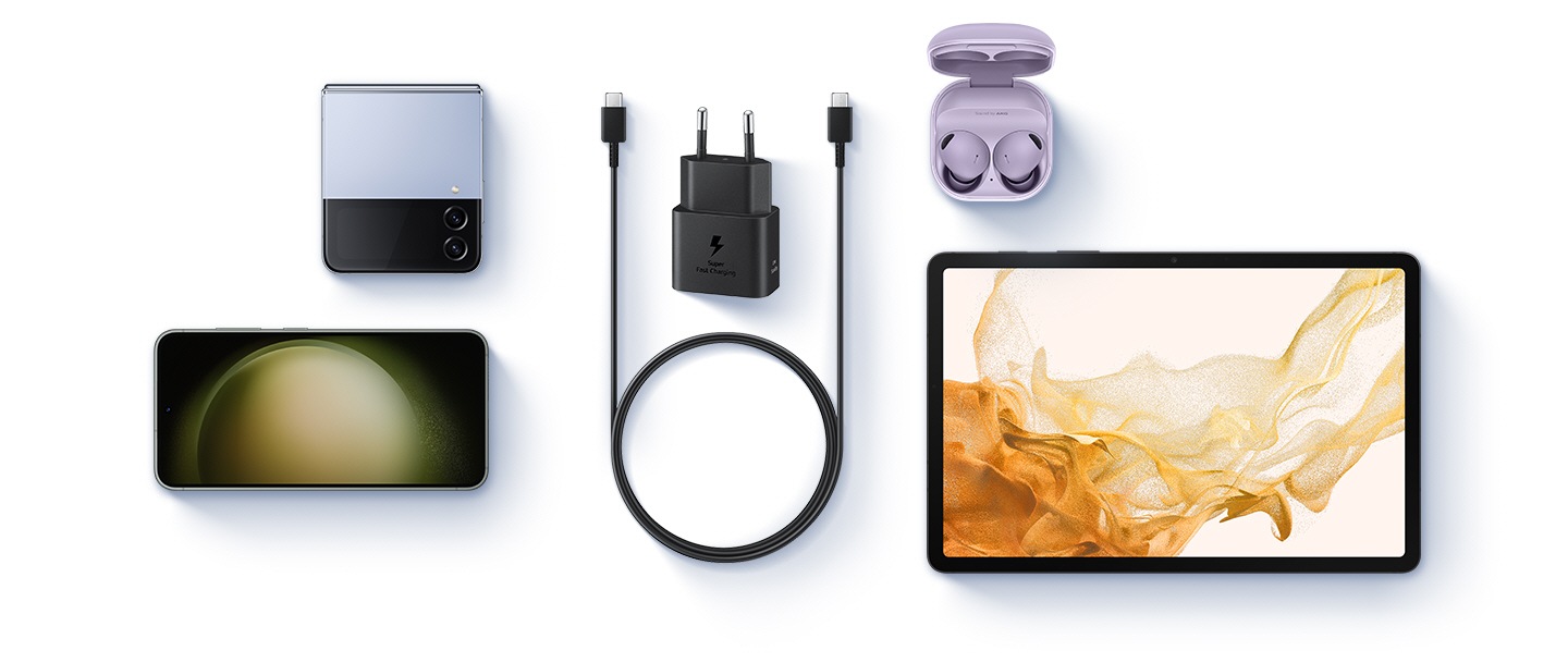 Pohled z ptačí perspektivy na různá zařízení je zobrazen s černým napájecím adaptérem a kabelem USB-C umístěným uprostřed. Vlevo jsou zobrazeny Blue Galaxy Z Flip4 a Galaxy S23. Napravo jsou zobrazeny Buds 2 Pro v barvě Bora Purple a Galaxy Tab S8.