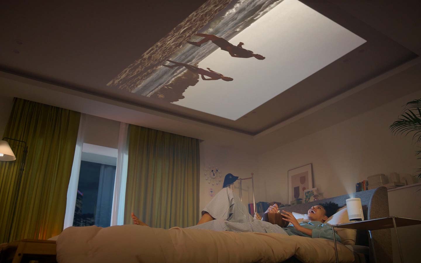 A woman is sitting on her bed eating popcorn, watching a video The Freestyle is projecting onto the wall in front of bed. Then she tilts The Freestyle so it points upwards and lays back with her head on the pillow, continuing to enjoying the video that’s now projected onto the ceiling.