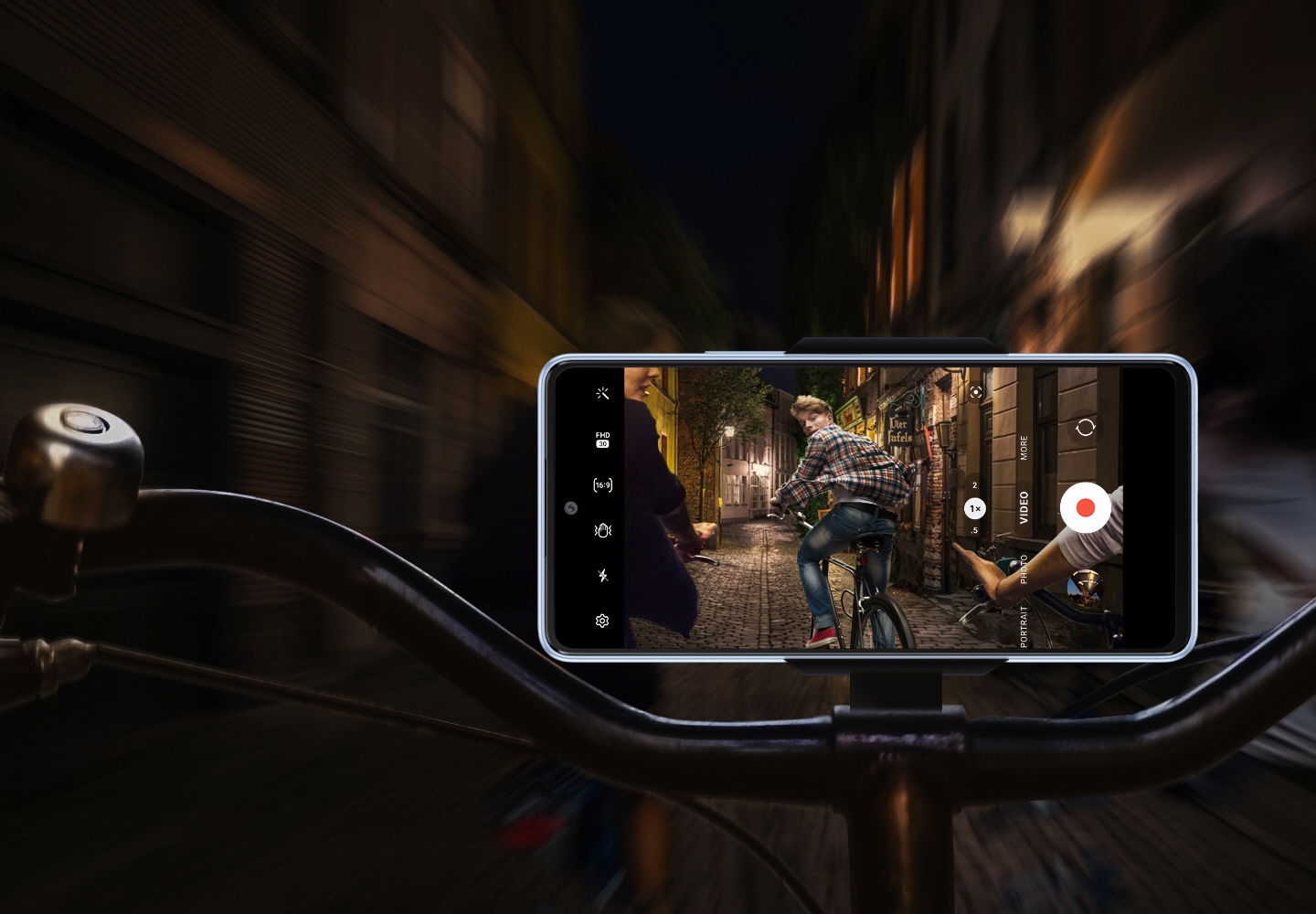 Frontalansicht eines Galaxy A53 5G, das waagerecht an einem Fahrradlenker befestigt ist. Während der Hintergrund unscharf und dunkel ist, zeigt das Smartphone-Display, dass es das Geschehen klar und stabil aufzeichnet: Eine Gruppe von Fahrradfahrern fährt nachts durch eine schwach beleuchtete Straße, wobei der Fahrer, der am weitesten vorne sitzt, auf das Smartphone schaut. 