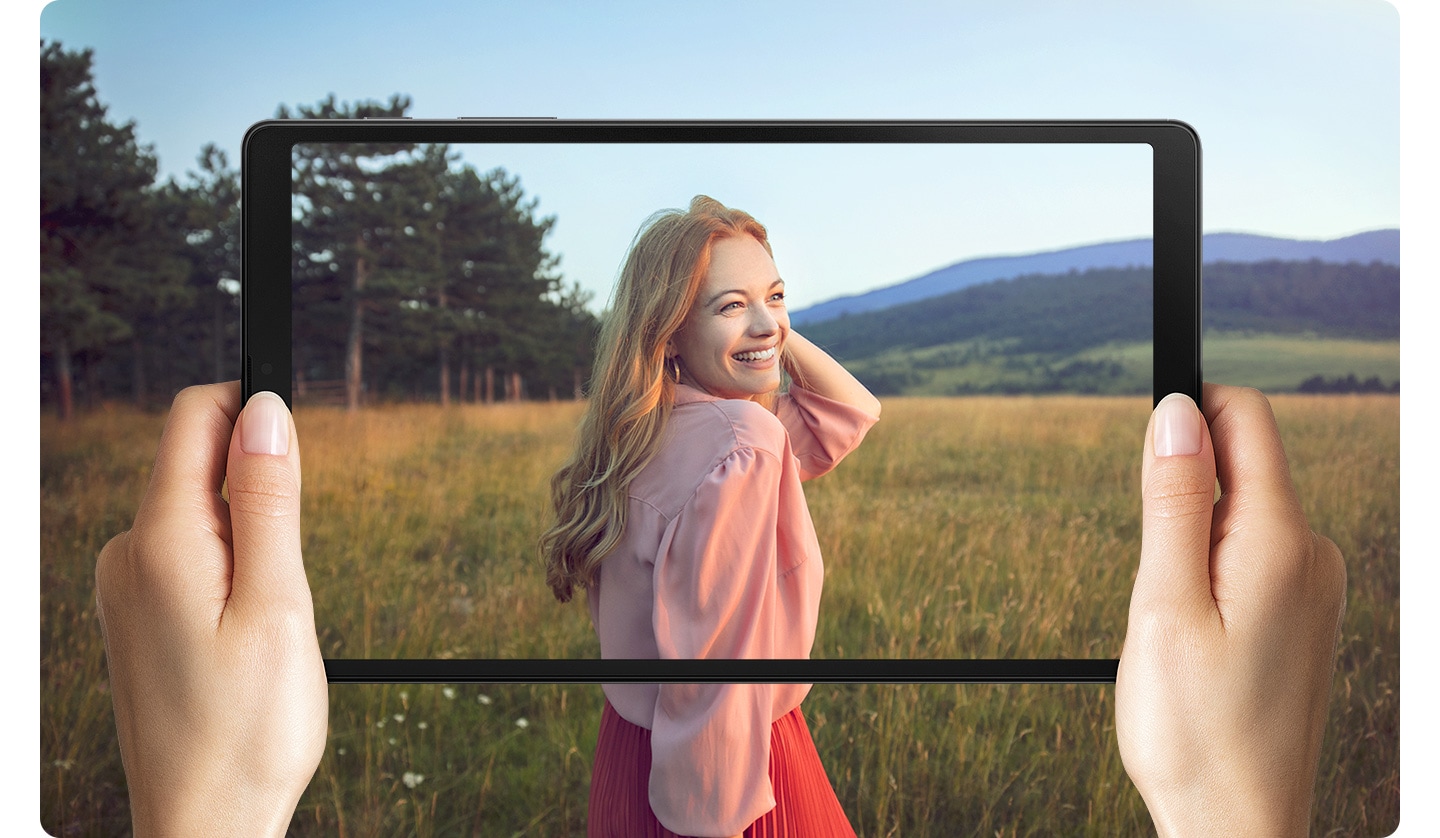 Hands holding Galaxy A7 Lite. Onscreen is a woman standing in a field. The photo goes beyond the boundaries of the screen to represent the wide display.