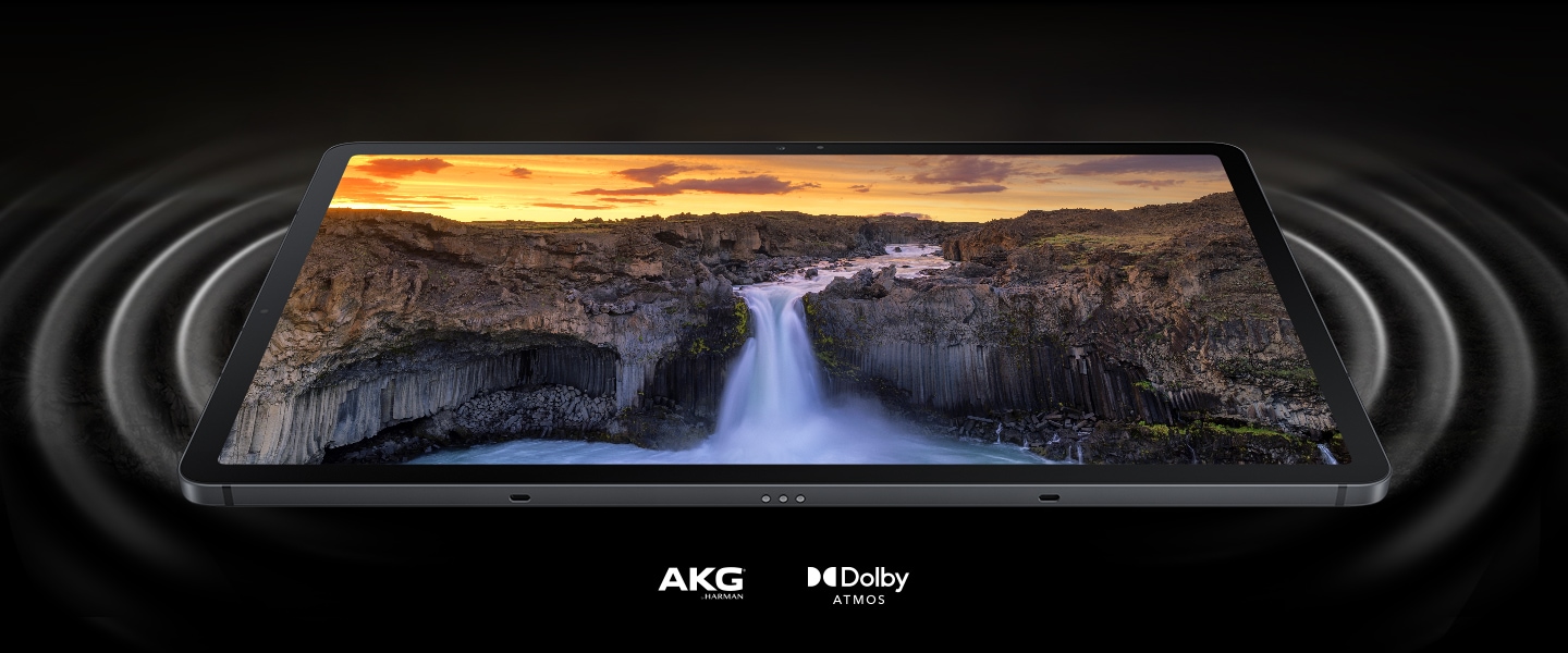 Das Galaxy Tab S7 FE 5G liegt auf der Rückseite und zeigt eine lebendige Landschaftsszene auf dem Bildschirm. Auf beiden Seiten des Tablets befinden sich Kreise, die Schallwellen darstellen, die aus den Dual-Lautsprechern kommen und den beeindruckenden Sound veranschaulichen. Man sieht AKG-Logo und Dolby Atmos-Logo.