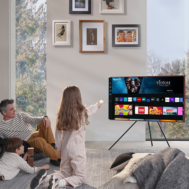 A girl is pointing the remote towards The Frame, which shows the Smart Hub home screen.