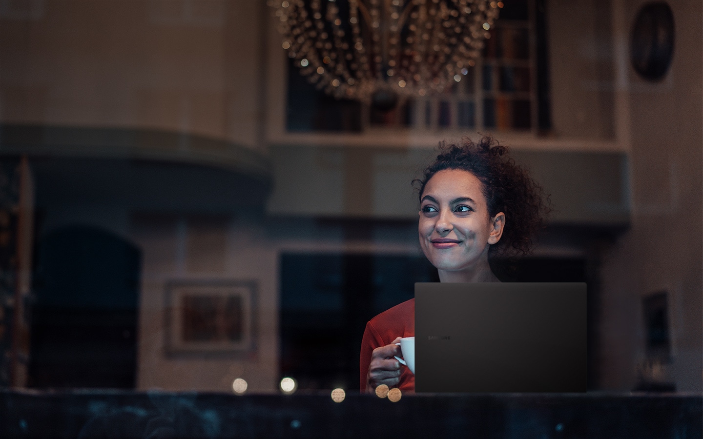 A woman is sitting down in a living room space that is luxuriously decorated with a chandelier and some artwork. As she holds a cup with her right hand behind Galaxy Book2 Pro, she is looking to the left, smiling.