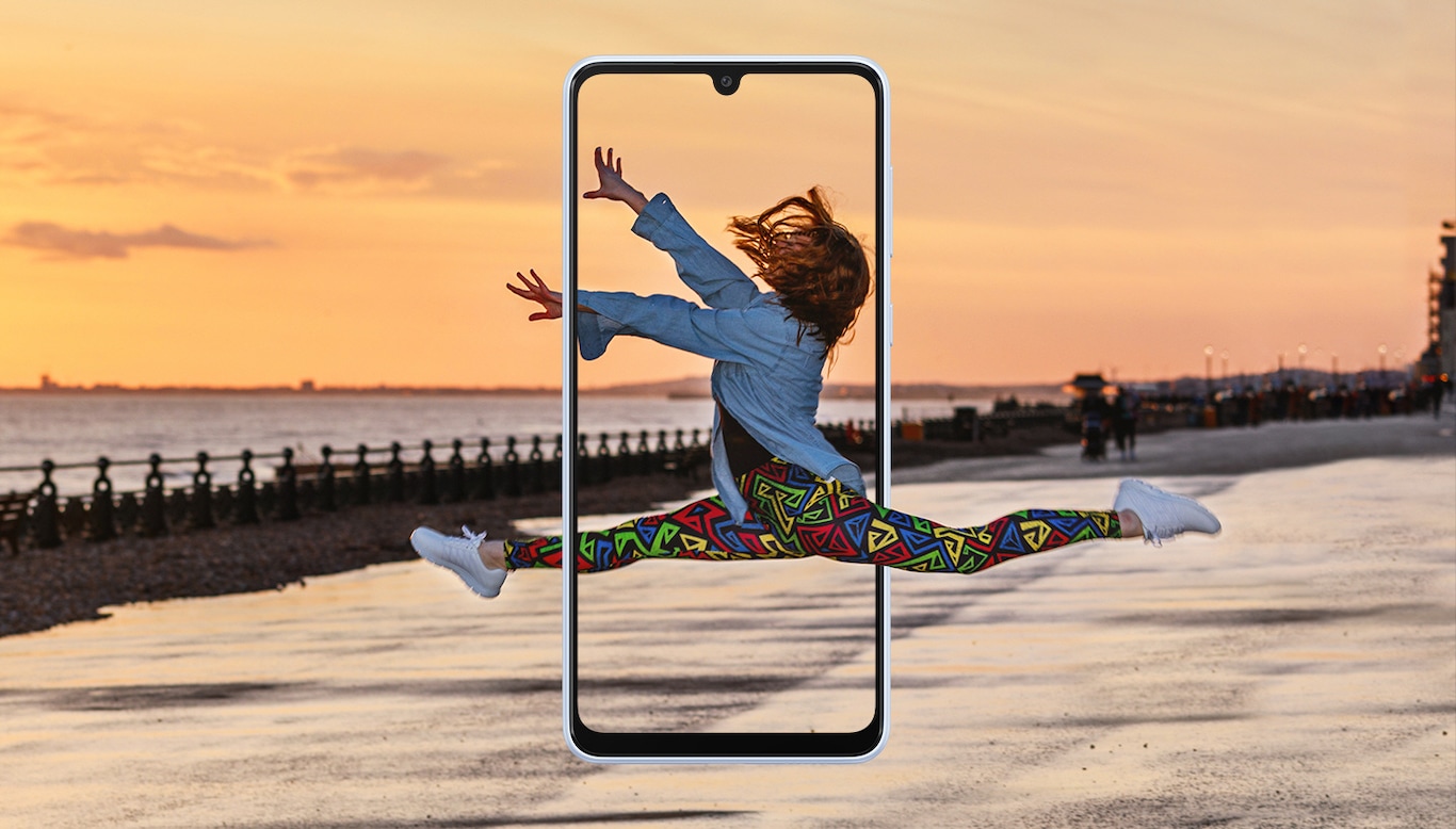 Eine Frau wird mitten im Sprung eingefangen, der an eine Ballettübung erinnert. Die Landschaft im Hintergrund zeigt einen wunderschönen Sonnenuntergang, und in der Mitte befindet sich ein Galaxy A33 5G-Bildschirm, der die Frau in der Mitte des Sprungs einrahmt.