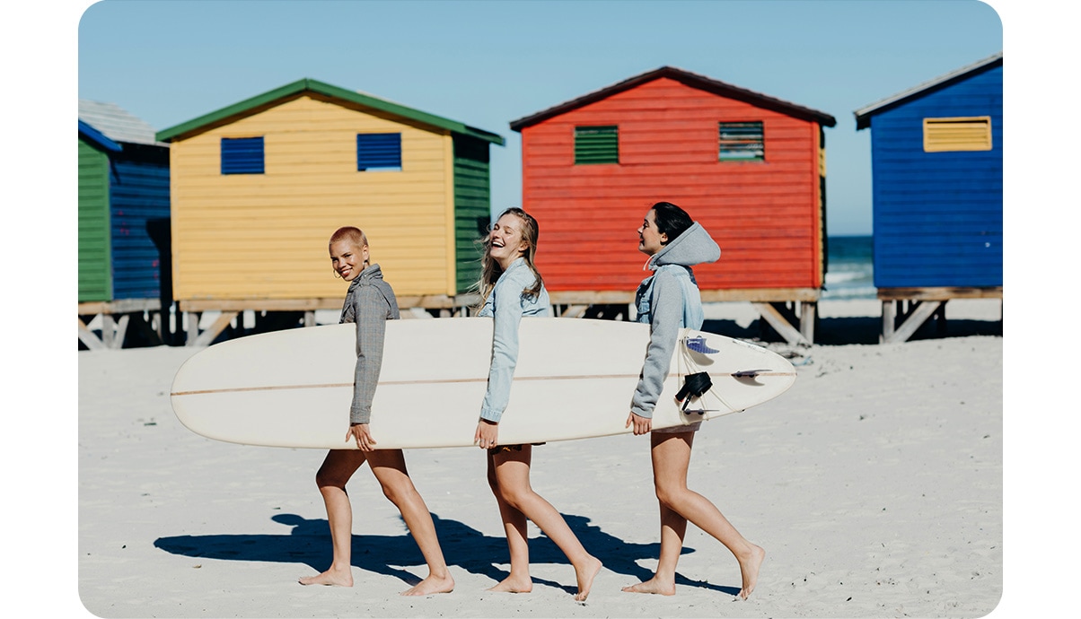 Drei lachende Frauen tragen ein Surfbrett, während sie einen Strand mit bunten Hütten im Hintergrund entlanglaufen. 