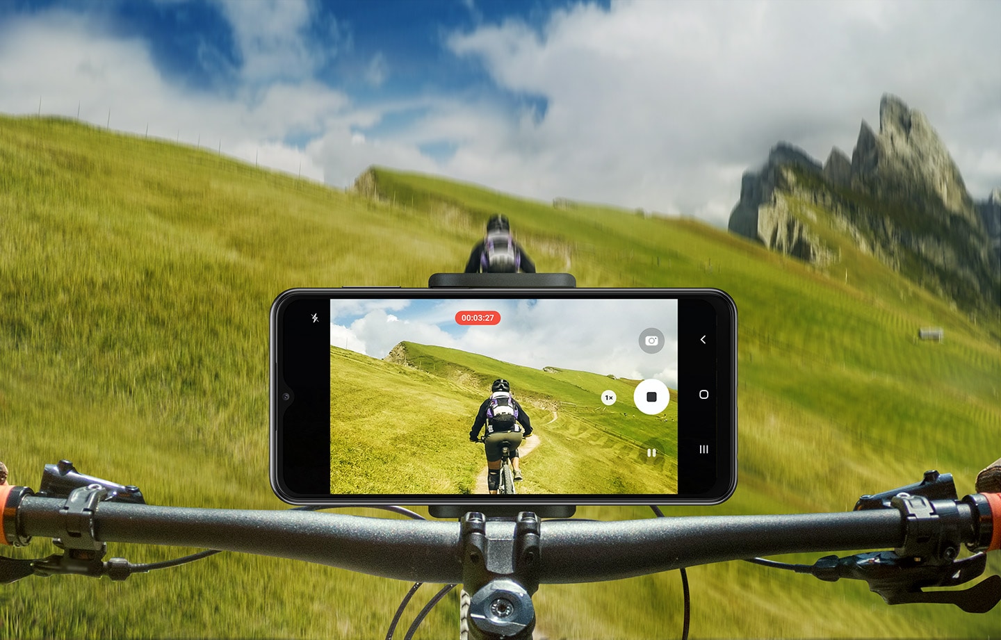 Ein Galaxy A23 5G ist am Lenker eines Mountainbikes befestigt, das im Gelände auf einer grasbewachsenen Piste gefahren wird. Die Kamera des Smartphones nimmt den Radfahrer vor ihm auf.