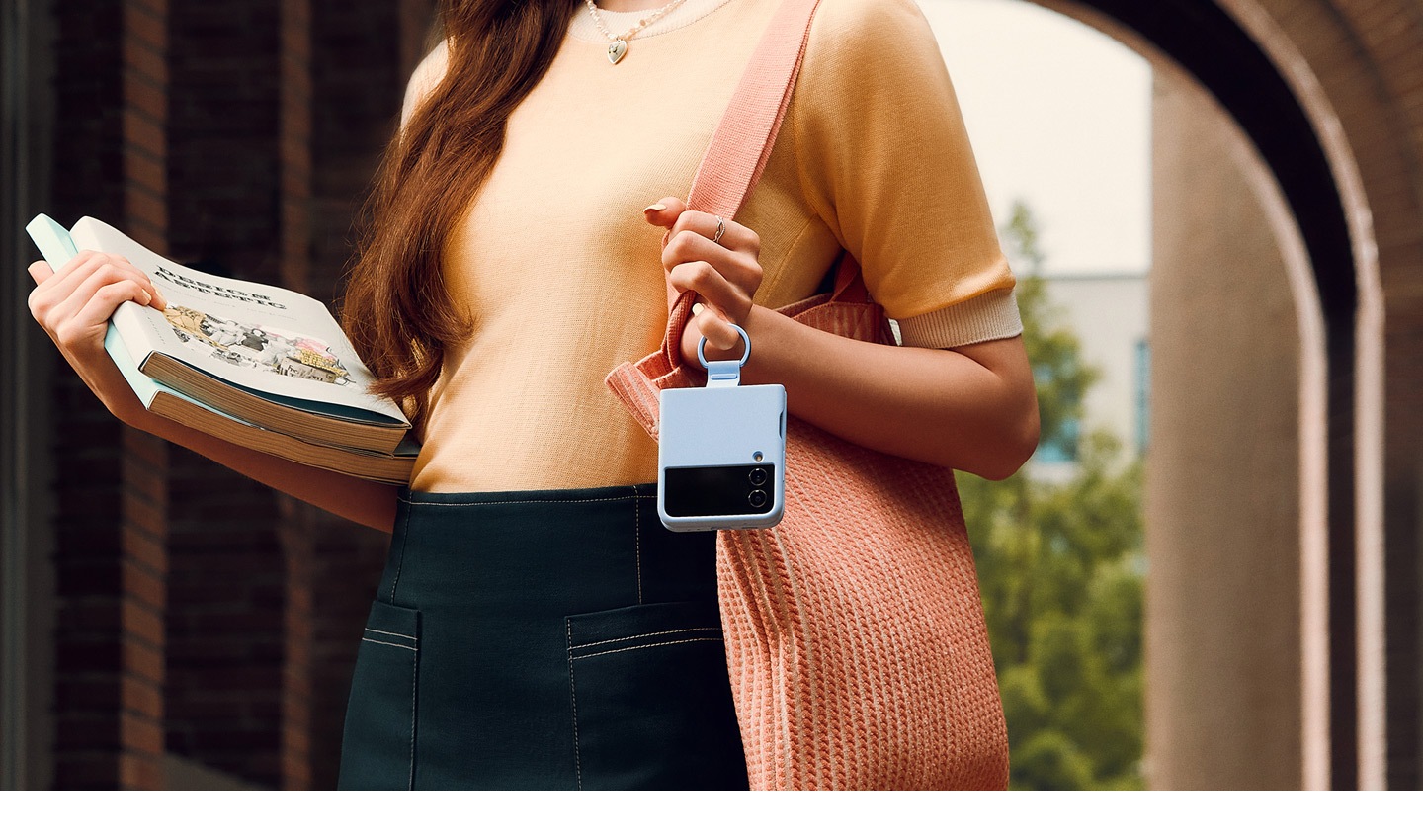 Eine Frau hält einen Stapel Bücher in einer Hand, während sie ihr Galaxy Z Flip4 in der anderen Hand am Ring eines Silicone Cover with Ring in Arctic Blue hält.