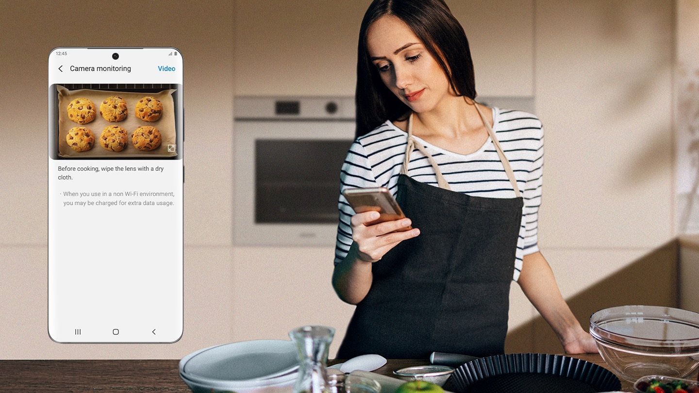 Shows a woman in front of an oven looking at her smartphone. A close-up of the phone screen shows the Camera Monitoring function of the SmartThings App, which is displaying a real-time video of cookies being baked in the oven.