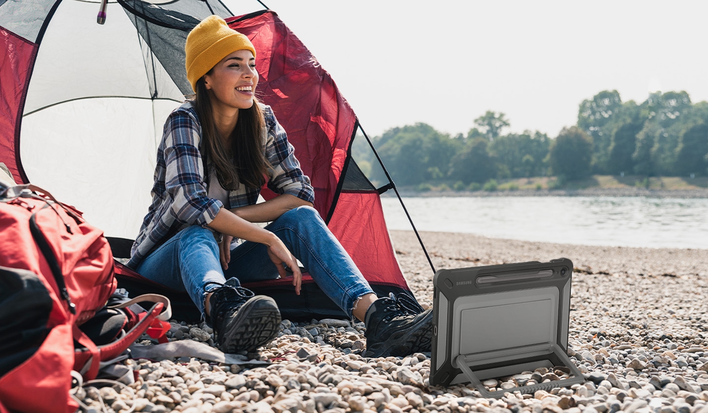 A woman sitting in front of a camping tent on a rocky surface is smiling. A Galaxy Tab S9 FE device with an Outdoor Cover on is placed horizontally in front of her propped up on a kickstand on the back of the cover.
