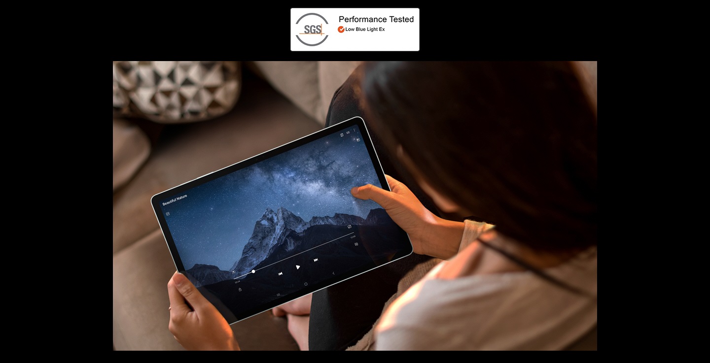 Eine Frau, die das Galaxy Tab S9 FE Plus mit beiden Händen auf einem Sofa hält und ein Video im Vollbildmodus im Querformat anschaut. SGS-Logo mit Text Performance Tested. Niedriges blaues Licht Ex.