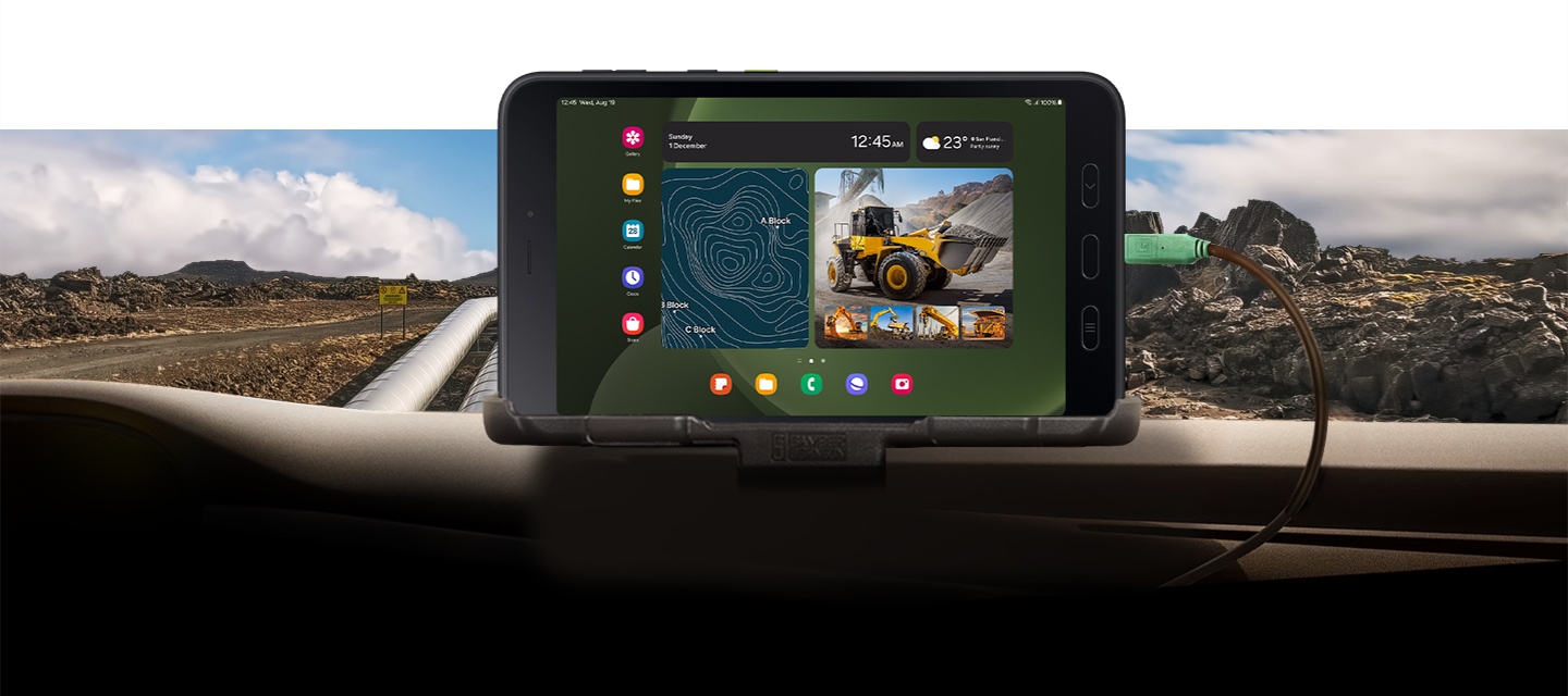 Ein Galaxy Tab Active5 5G ist auf dem Armaturenbrett eines Autos montiert und zeigt auf dem Bildschirm eine GPS-Karte und Bilder von Fahrzeugen mit schwerem Gerät. Das Gerät ist über ein Kabel mit dem Auto verbunden, und im Hintergrund ist ein unwegsames, felsiges Gelände zu sehen.