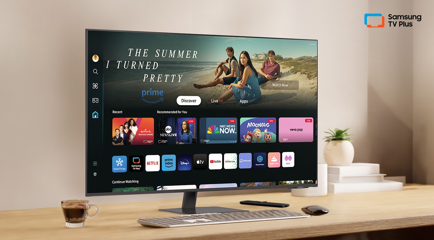 There is a monitor on a desk and its home UI is shown with OTT applications. On the upper right, there's a logo of Samsung TV Plus.