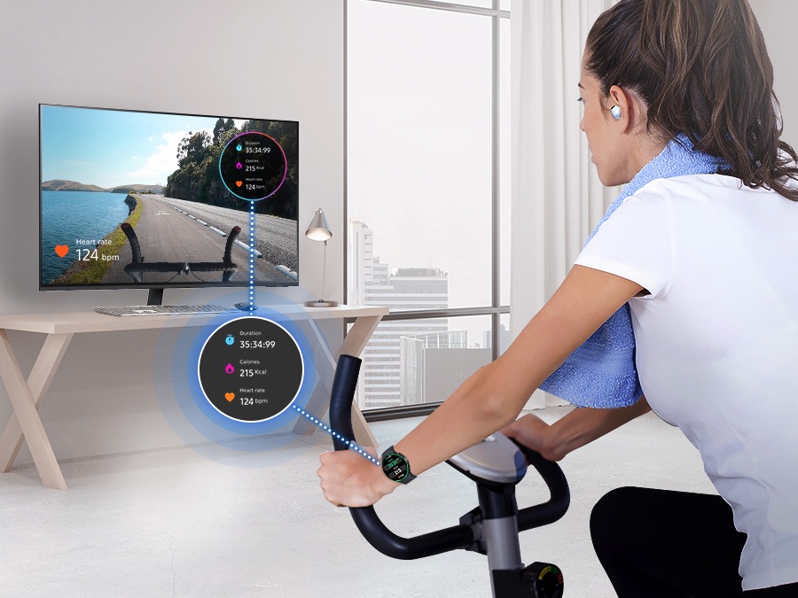 A woman is riding an indoor bicycle. She is wearing a Galaxy Watch and Buds, and her watch screen is overlaid on the monitor.