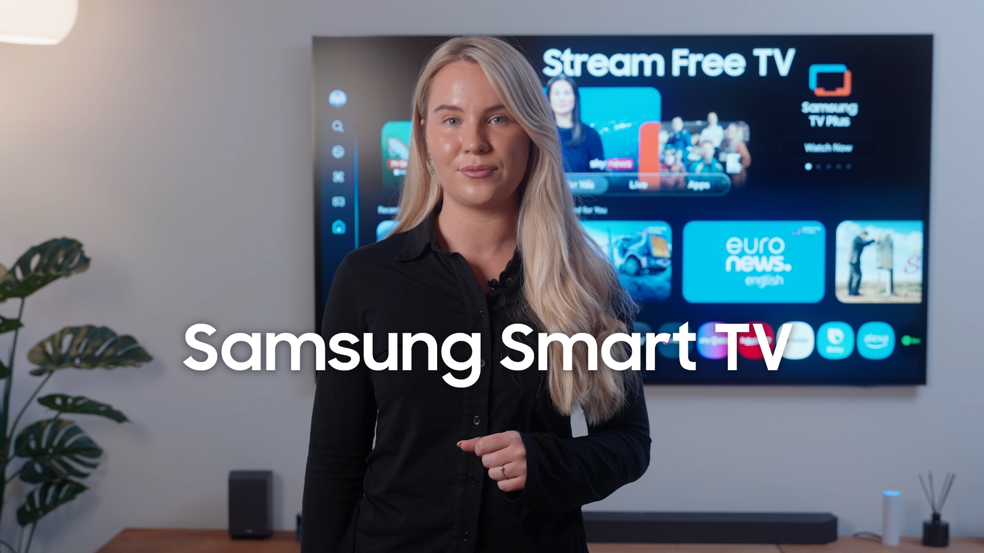 En person står foran et Samsung Smart-TV, der viser brugerfladen til gratis streaming. Skærmen viser apps som "Samsung TV Plus" og "euronews english" samt videominiaturer. I baggrunden ses en lampe, en plante og en soundbar i en moderne stue.