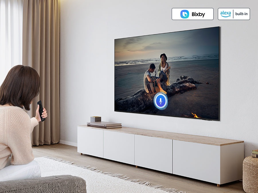 Bir kadın televizyonunu kontrol etmek için sesli asistanını kullanıyor. "Bixby" ve "Alexa yerleşik" logoları.