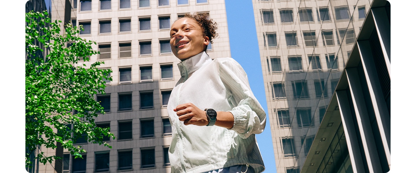 A woman in sportswear wearing a Galaxy Watch7 walks along a city block surrounded by other buildings.