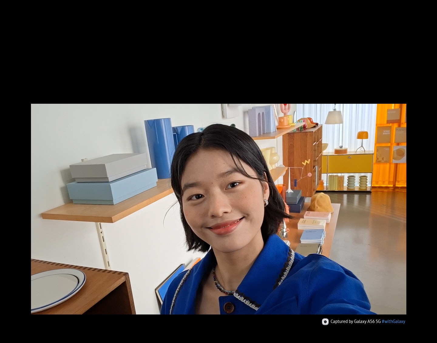 Person in a showroom, smiling. The person turns and grabs a cup from a shelf. The person turns back to face the front, smiling with the cup next to the face. At the bottom is camera icon with text Captured by Galaxy A56 5G hashtag with Galaxy.