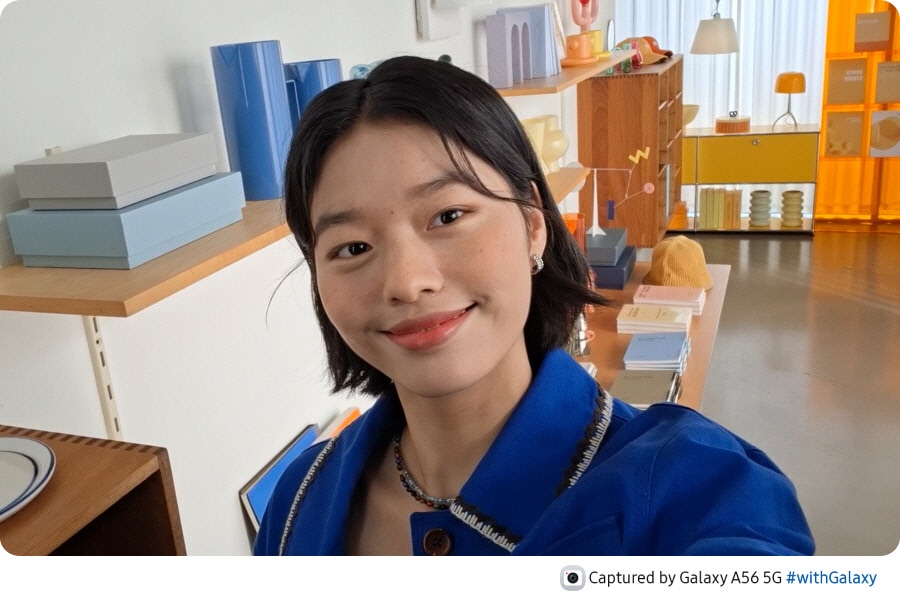 Person in a showroom, smiling. The person turns and grabs a cup from a shelf. The person turns back to face the front, smiling with the cup next to the face. At the bottom is camera icon with text Captured by Galaxy A56 5G hashtag with Galaxy.