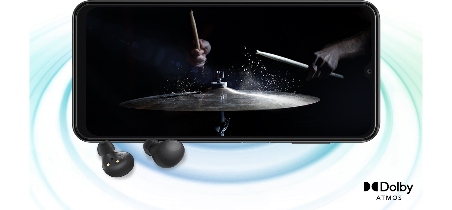 Galaxy A13 in landscape mode and an image with a person playing drums in the black background onscreen. A pair of black Galaxy Buds2 are placed in front of the device. On the right bottom is a logo for Dolby Atmos.