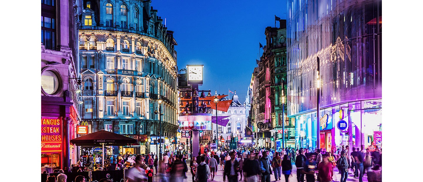 A busy city center with people walking around is very dimly lit. A box zooms into the clock which then shows a comparison of the image with FHD and UHD. A wave of pixels then rushes over the scene and creates a more vivid, clearer video quality in UHD.