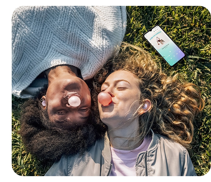 Two female friends, both wearing Buds Pro, are lying down on the grass on a sunny day, blowing bubbles with bubble gum. A smartphone laid down nearby shows a song being played, illustrating that both friends are listening to the song together with their Buds products.