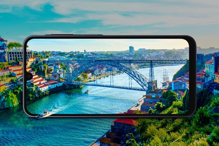 An illustration of a cityscape with a river running through and a bridge connecting two sides. A large screen of Galaxy A16 in the center displays a real photo of the scene. A boat on the river appears as if coming out of the screen, showcasing the device's immersive display quality.