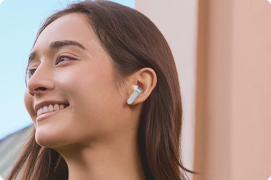 La vista esquelética de los Galaxy Buds4 está en el centro. A medida que cambian las formas de las orejas y las condiciones de uso, los Galaxy Buds4 se ensamblan. Los auriculares encajan en la oreja de una persona, quien sonríe y mira hacia la izquierda.