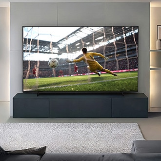 A large TV in a modern living room shows a soccer goalie diving to save a goal in a stadium, with a gray sofa and rug visible.